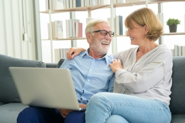 Casal idoso sorridente sentado no sofá, olhando um para o outro com carinho, enquanto um deles segura um laptop no colo. Ao fundo, uma estante de livros.